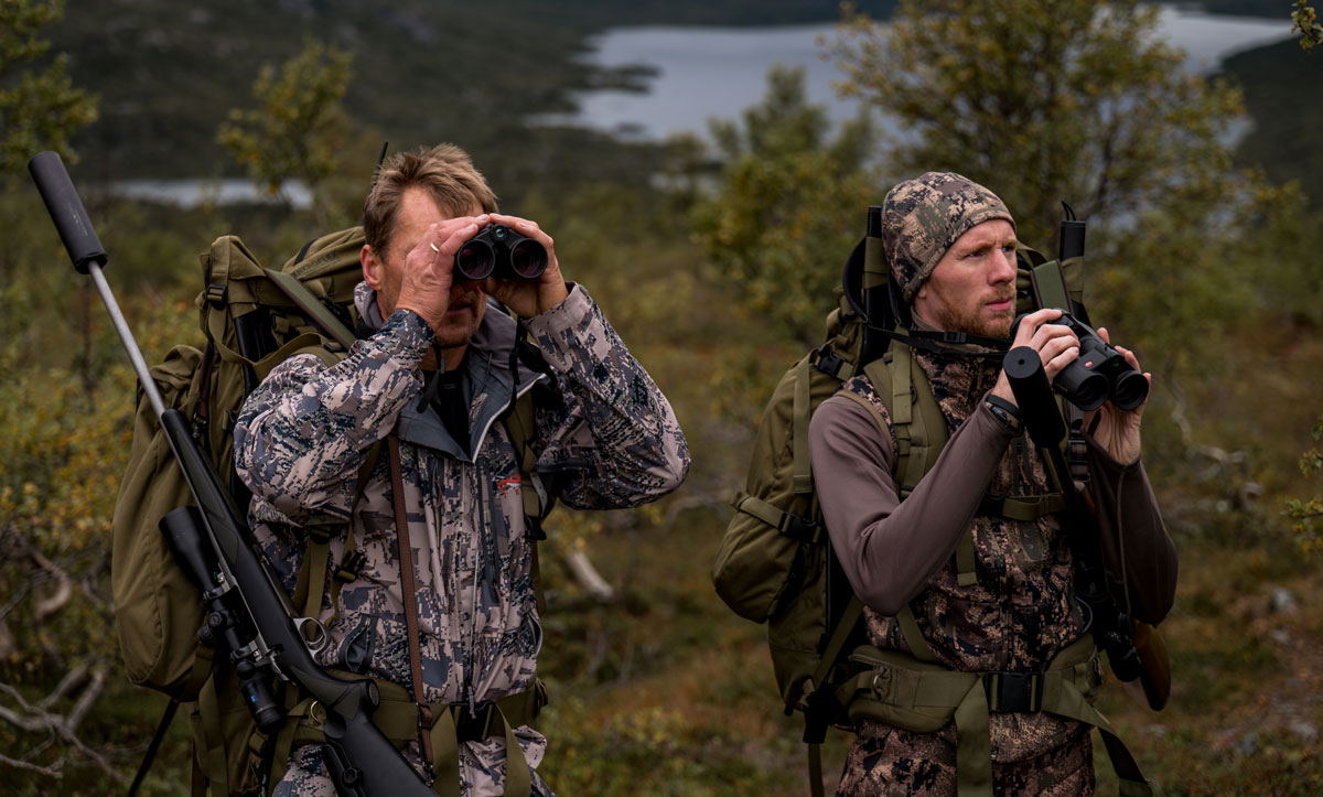 Two hunters in camouflage clothing go through a rocky landscape with backpacks
